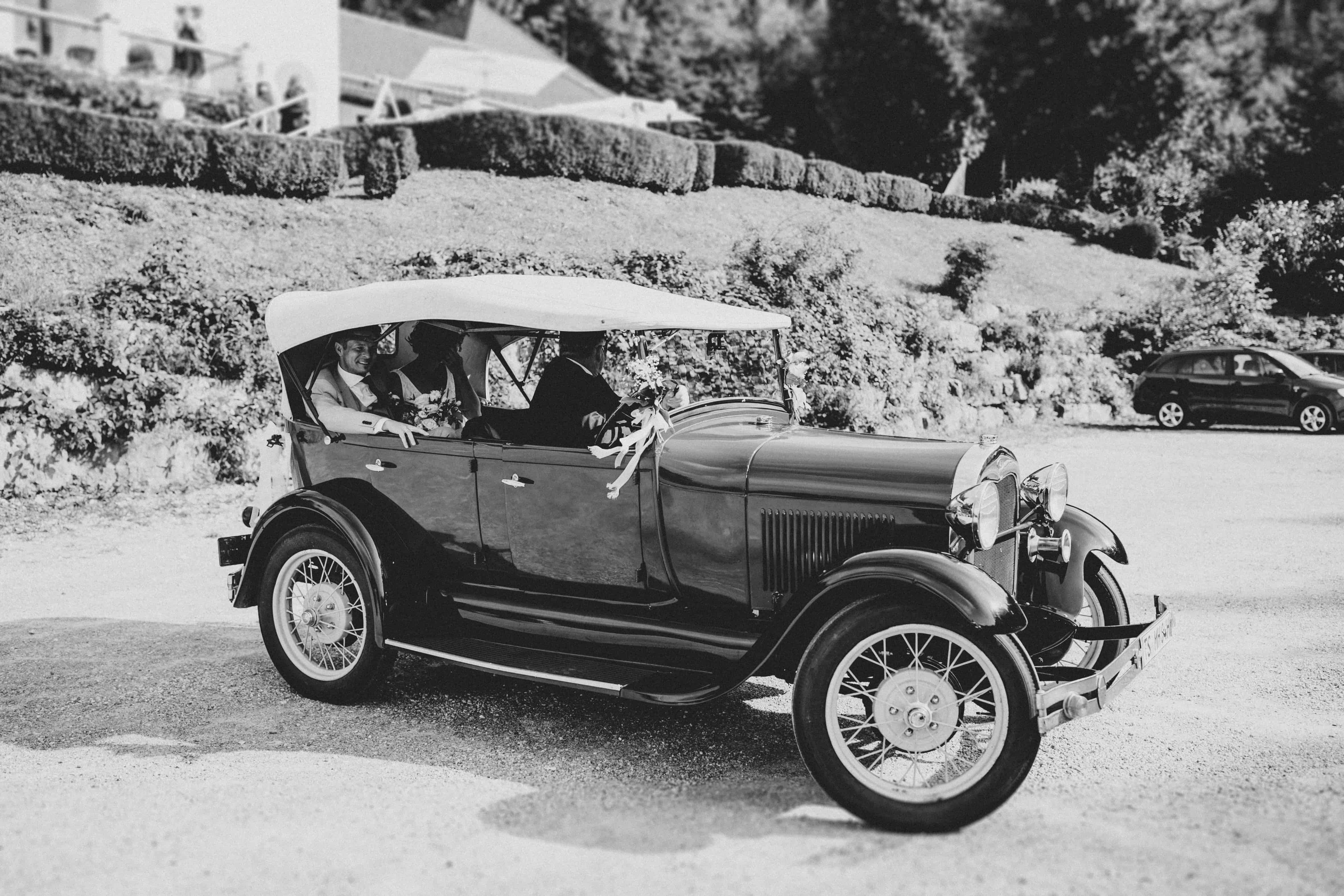 Oldtimer Brautauto mit frisch vermählten Brautpaar und Chauffeur beim Parken bei der Hochzeitslocation bei einer wunderschönen Sommerhochzeit in den Bergen mit Hochzeitsplanerin pure passion weddings by Anna Walch