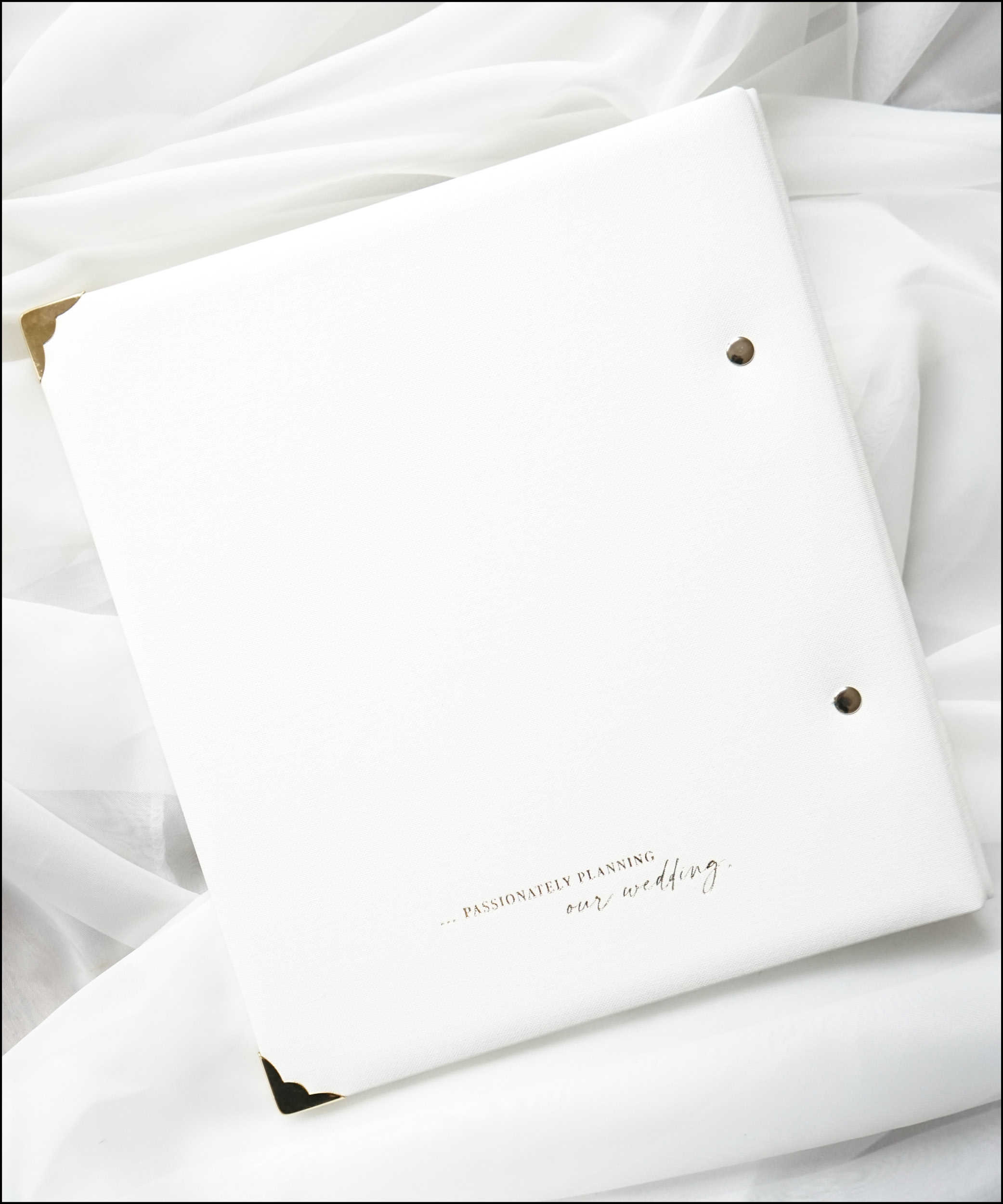 Dieses Bild zeigt das Buchcover-Rückseite des Crème-de-la-Crème DIY Hochzeitsplaners. Es zeigt die Goldprägung "...passionately planning our wedding" auf dem hochwertigen, gepolsterten, weißen Leinen-Cover und die goldenen Buchecken zum Schutz dieses Premium Hochzeitsplaners.